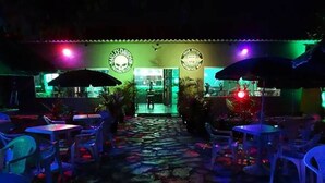 An outdoor patio area with tables and chairs, a stone pathway, and a building with neon signage in the background. - Road House GO 118 Motorcycle Hotel (Alto Paraíso de Goiás)