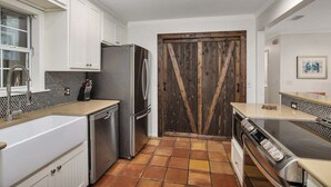 Eine Küche mit weißen Schränken, einem Edelstahlkühlschrank, einer Holztür und einem Fliesenboden.