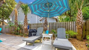 Ein Garten mit einer Terrassenfläche, inklusive zwei Liegestühlen und einem Tisch, einem gestreiften Sonnenschirm und einem Holzzaun.