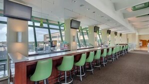 Uma área de bar moderna com um balcão comprido, banquetas verdes e amplas janelas com vista para o exterior.