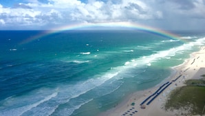 Uma praia com um arco-íris visível sobre o oceano, areal arenoso e algumas pessoas e guarda-sóis.