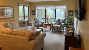 Un salon avec un canapé beige, deux fauteuils, une table basse et un téléviseur fixé au mur.