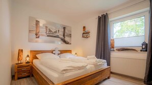 Un lit en bois avec du linge de lit blanc, une table de chevet avec une lampe, un tableau au-dessus du lit, une fenêtre avec des rideaux et un radiateur.