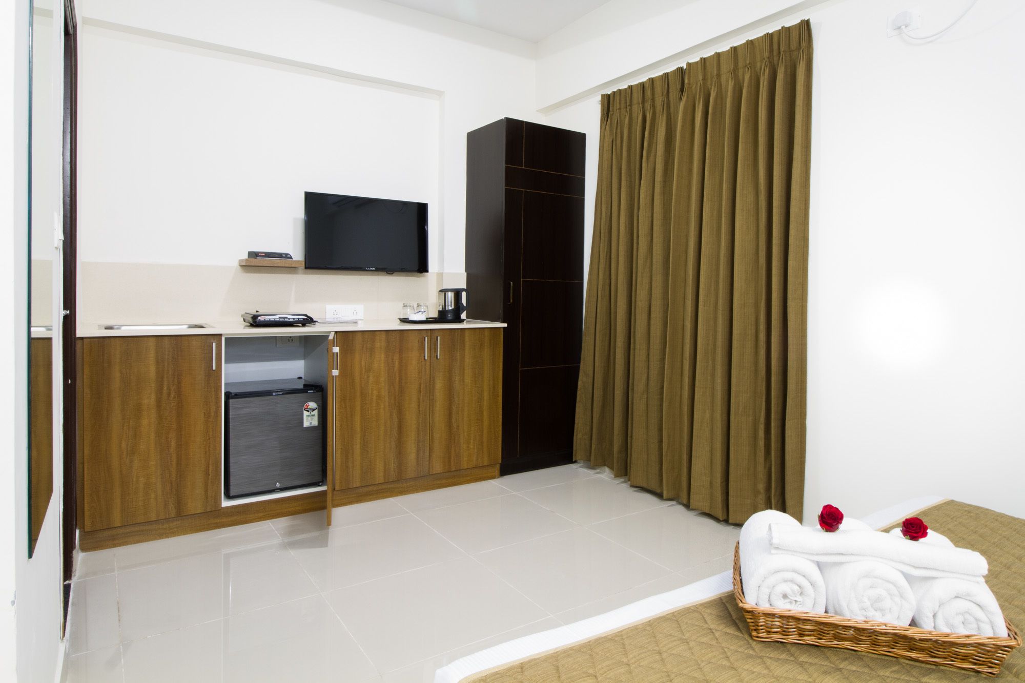 Una habitación de hotel moderna con kitchenette, una televisión de pantalla plana, un ventanal grande con cortinas, y una cesta de mimbre con toallas y rosas.