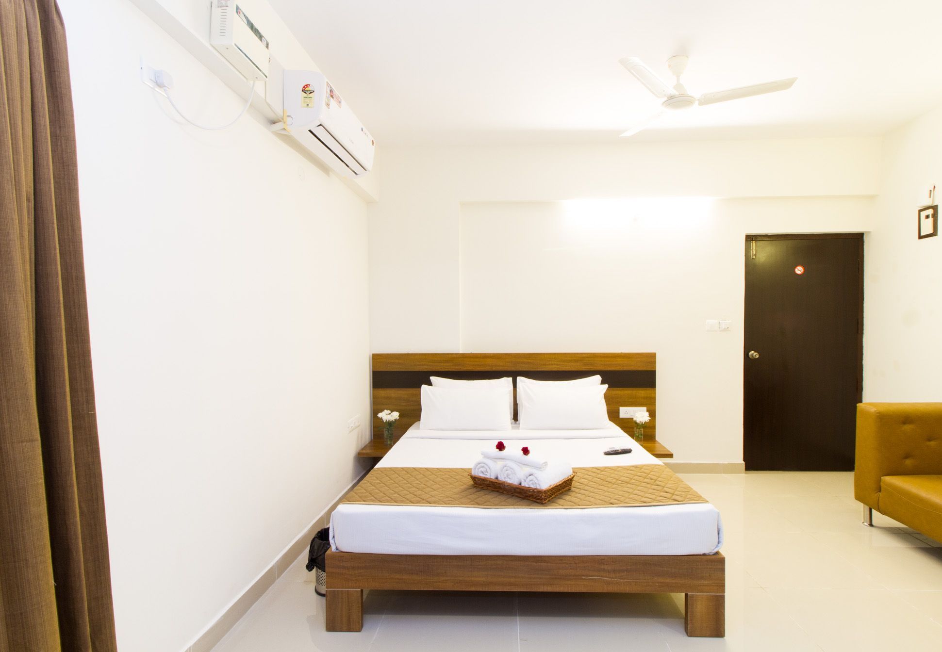 Une chambre d’hôtel avec un lit en bois, du linge de lit blanc, une petite table avec un panier, un ventilateur de plafond et une porte sur laquelle est apposé un panneau « interdiction de fumer ».