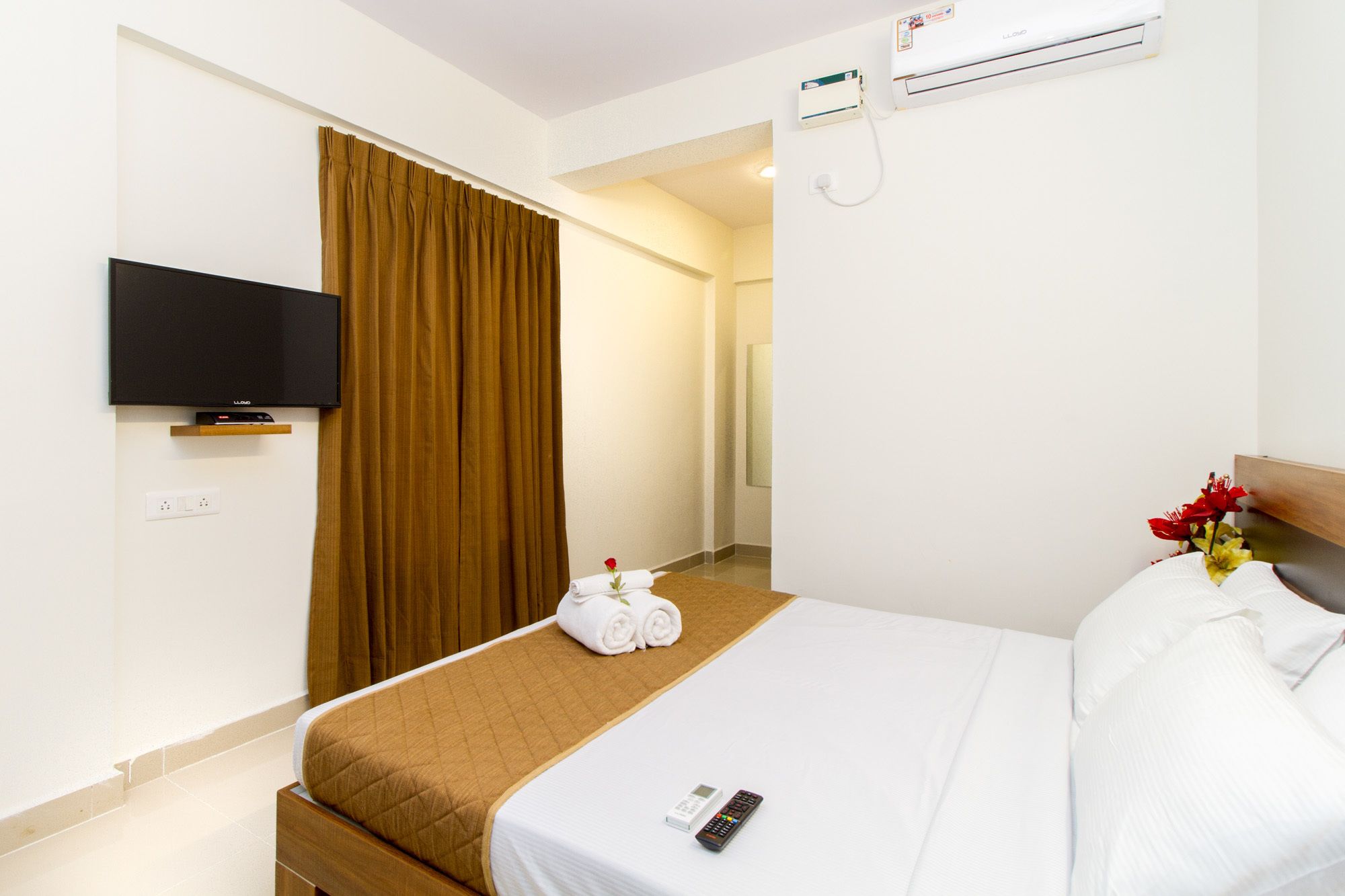 A hotel room with a bed, a TV mounted on the wall, an air conditioning unit, and a remote control on the bed.