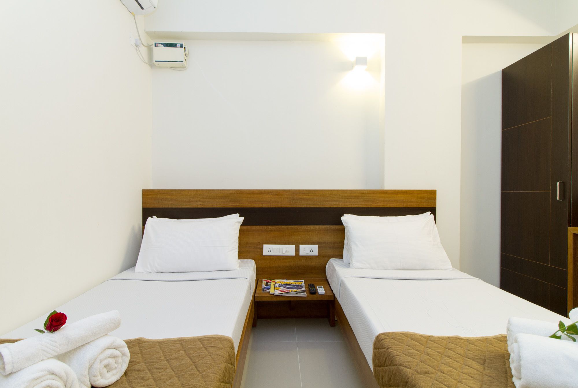 Une chambre d’hôtel avec deux lits simples, une tête de lit en bois, du linge de lit blanc, une table de chevet avec un magazine et une rose rouge sur le lit de gauche.