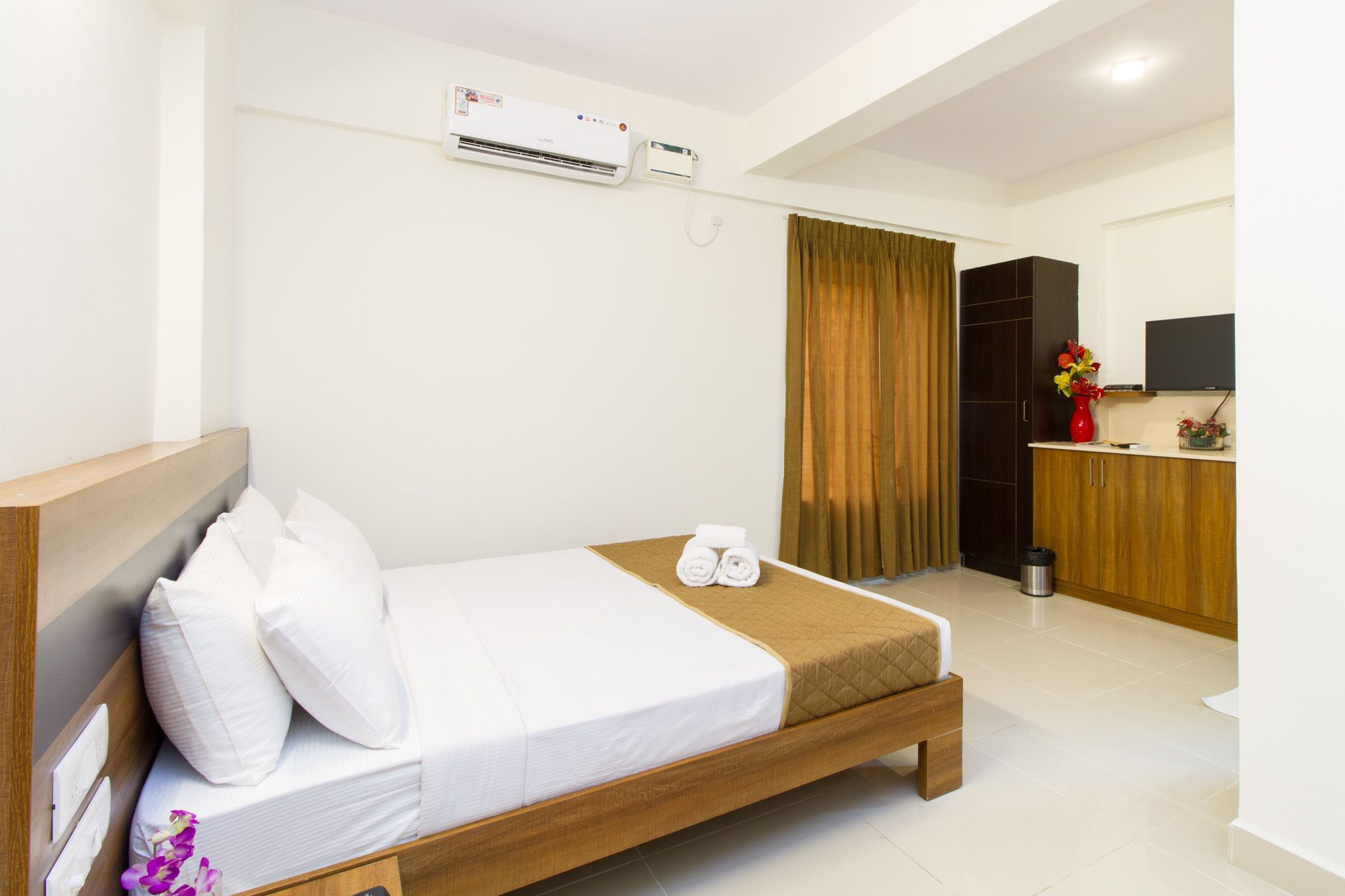 Une chambre d’hôtel comprenant un lit, une tête de lit en bois, une table de chevet, un téléviseur, un petit coin cuisine et un appareil de climatisation.