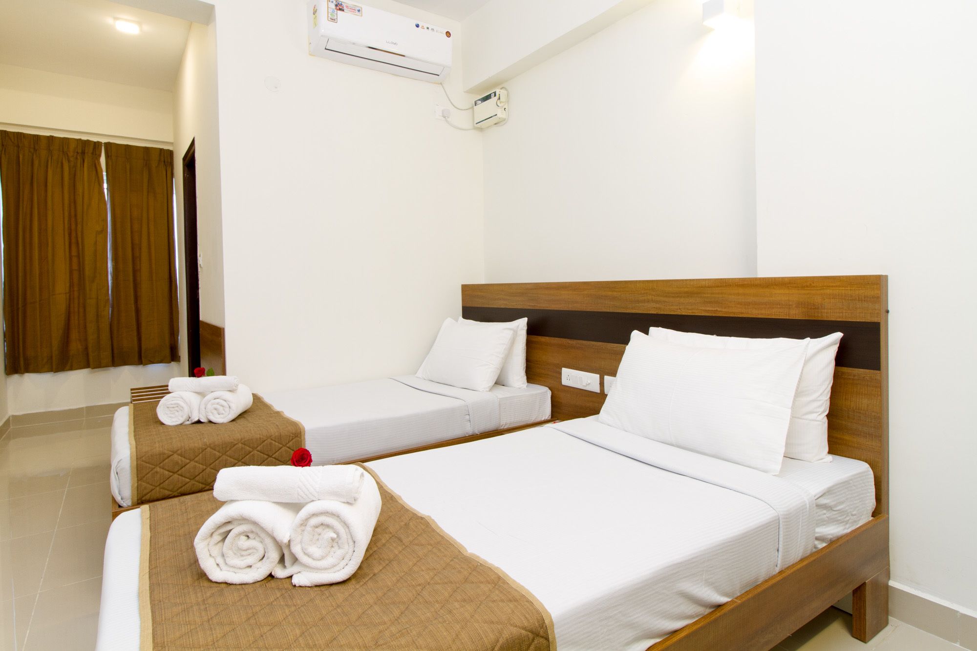 A hotel room with two beds, each with white linens and pillows, a wooden headboard, and a brown quilted bed runner. There is a rolled towel on the floor and a red rose on the bed runner.