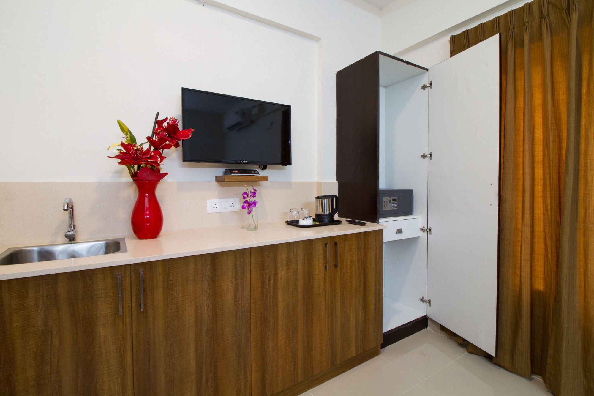 A modern kitchen with a wooden cabinet, a stainless steel sink, a red vase with flowers, a black television, and a coffee maker.