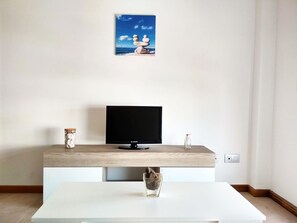 Una sala de estar moderna con un televisor de pantalla plana sobre un mueble de madera, un florero de vidrio con una planta y un cuadro enmarcado con una escena de playa en la pared.