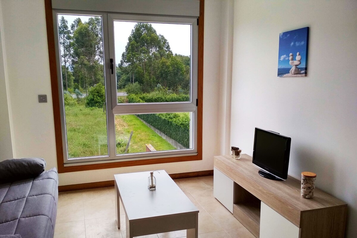 Una sala de estar moderna con un ventanal grande, un televisor sobre un mueble de madera, una mesita pequeña y un sofá gris.