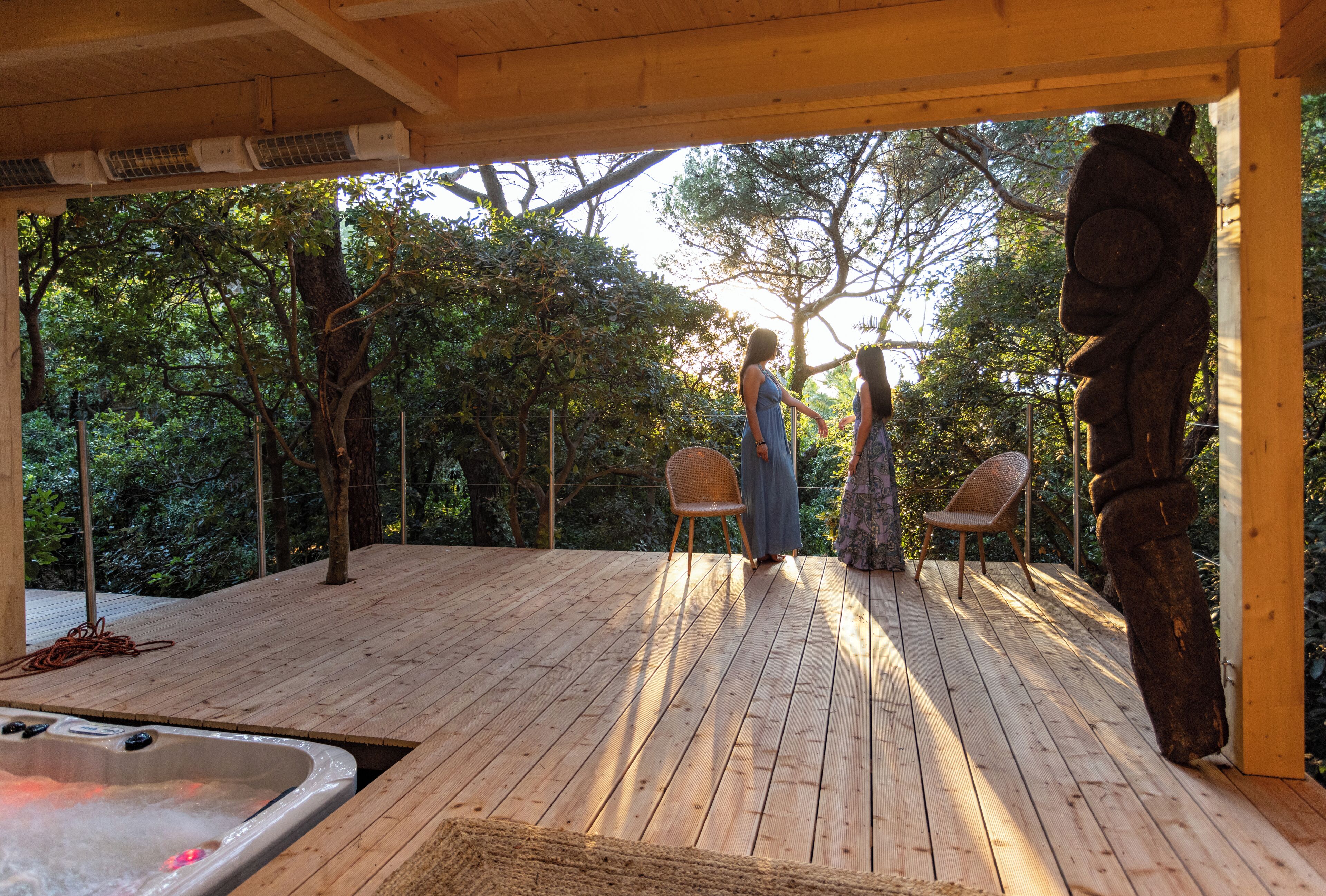 Een houten terras met een jacuzzi, twee rieten stoelen en een sculptuur.