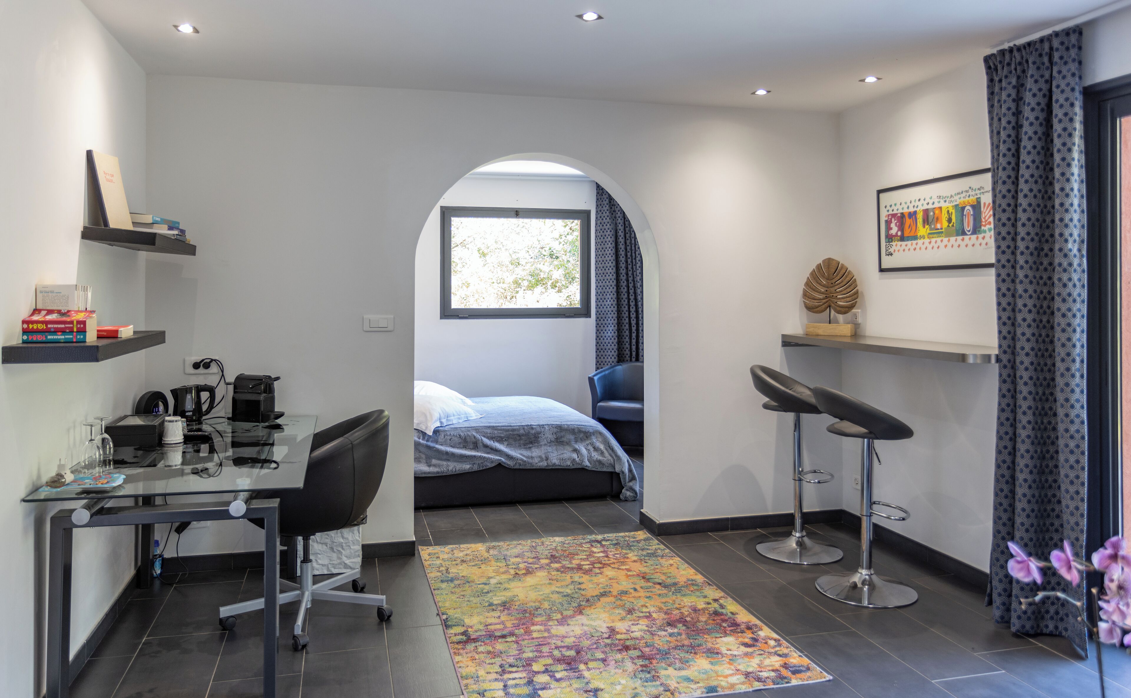 Un dormitorio moderno con cama, escritorio, silla y un pequeño rincón de descanso con dos taburetes.