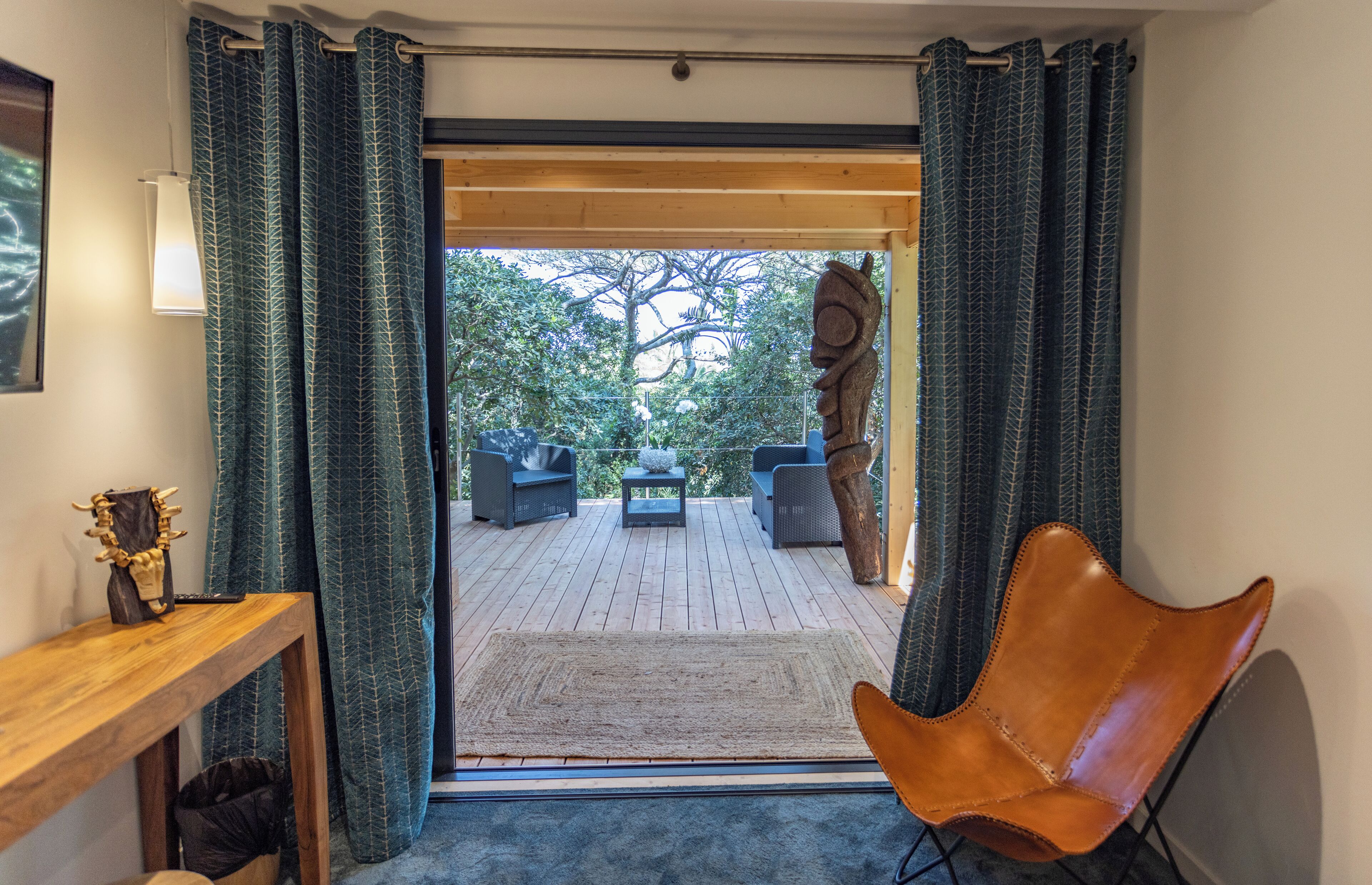 Vista desde el interior de una habitación a un balcón exterior con una mesa de madera, una silla y una escultura.