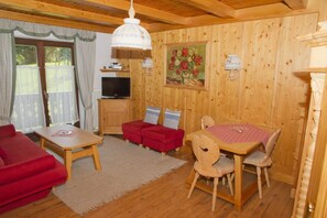 A cozy room with a wooden interior, a dining table with chairs, a red sofa, a television, and a painting of flowers. - Apartment Römerweg - House Römerweg (Strobl)