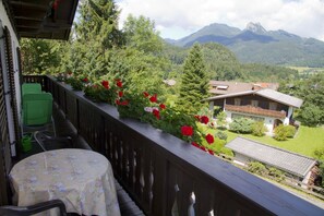 A balcony with a table and chairs, a flower border, and a view of a house and mountains. - Apartment Römerweg - House Römerweg (Strobl)