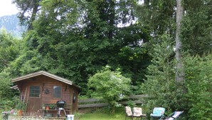 A rustic wooden cabin with a porch, surrounded by lush greenery and a wooden fence.