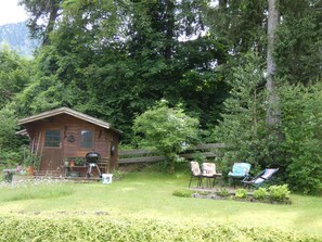 A rustic wooden cabin with a porch, surrounded by lush greenery and a wooden fence.