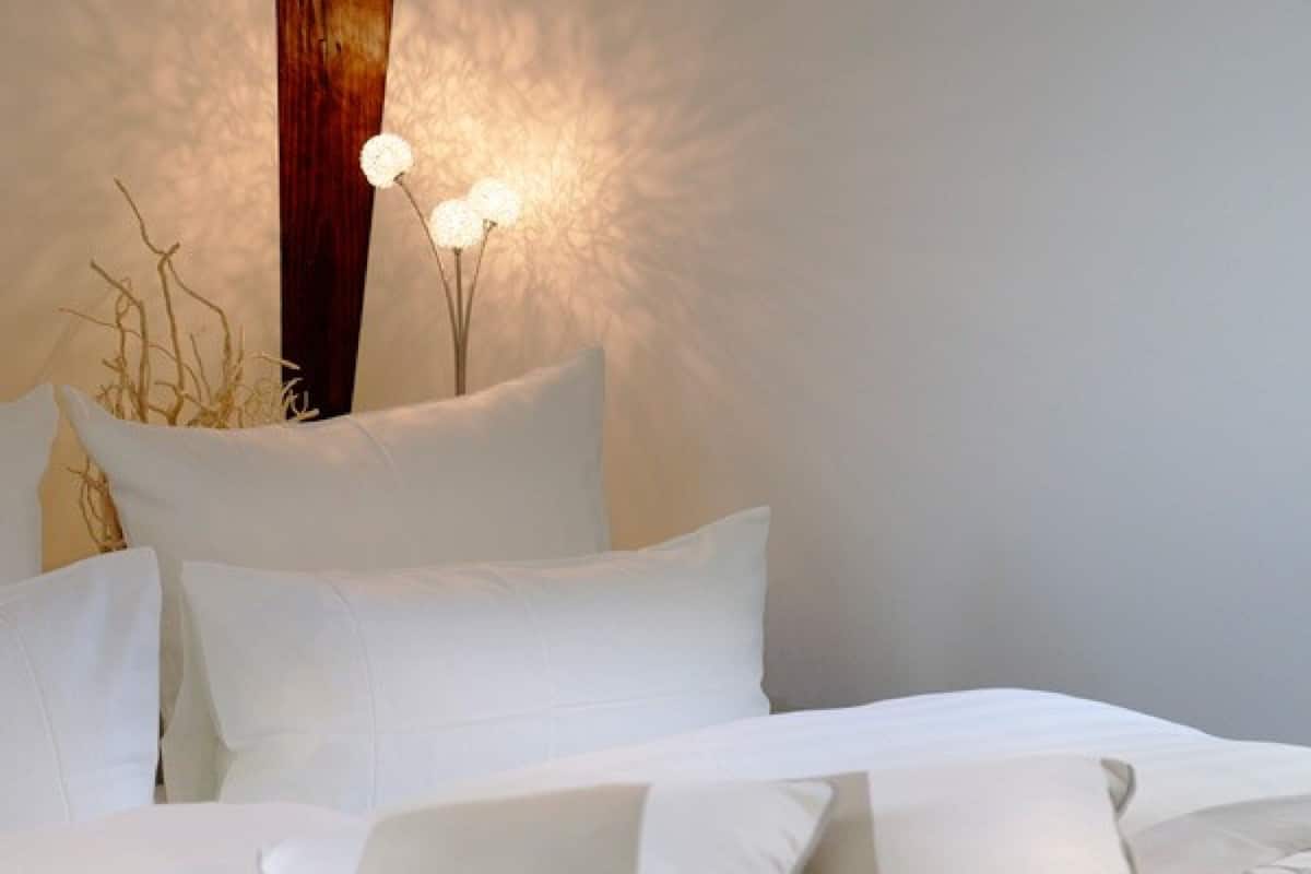 A neatly made bed with white bedding and pillows, a decorative wall with a wooden panel and wall-mounted lights, and a vase with dried branches.