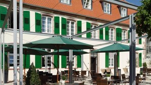 An outdoor cafe with tables and chairs under green umbrellas, set against a building with red-tiled roof and green shutters. - Schlosshotel Bergzaberner Hof (Bad Bergzabern)