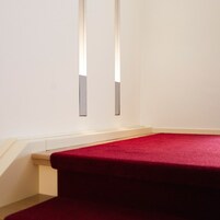 Esquina de una habitación con piso cubierto de alfombra roja y una pared blanca con dos lámparas verticales.