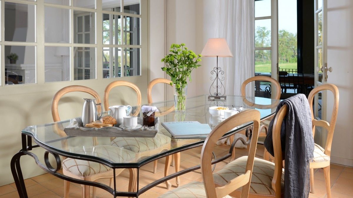 A dining area with a glass table, wooden chairs, a vase with green plants, a lamp, and a view of the outdoors through large windows.