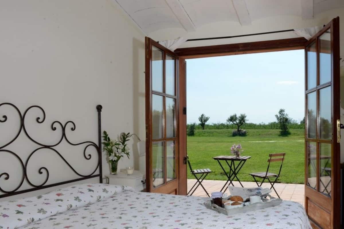 Een slaapkamer met een bed, een klein tafeltje met een dienblad eten, en uitzicht op een grasveld door open deuren.