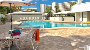 Una piscina con sillas de descanso y sombrillas, rodeada de edificios y palmeras.