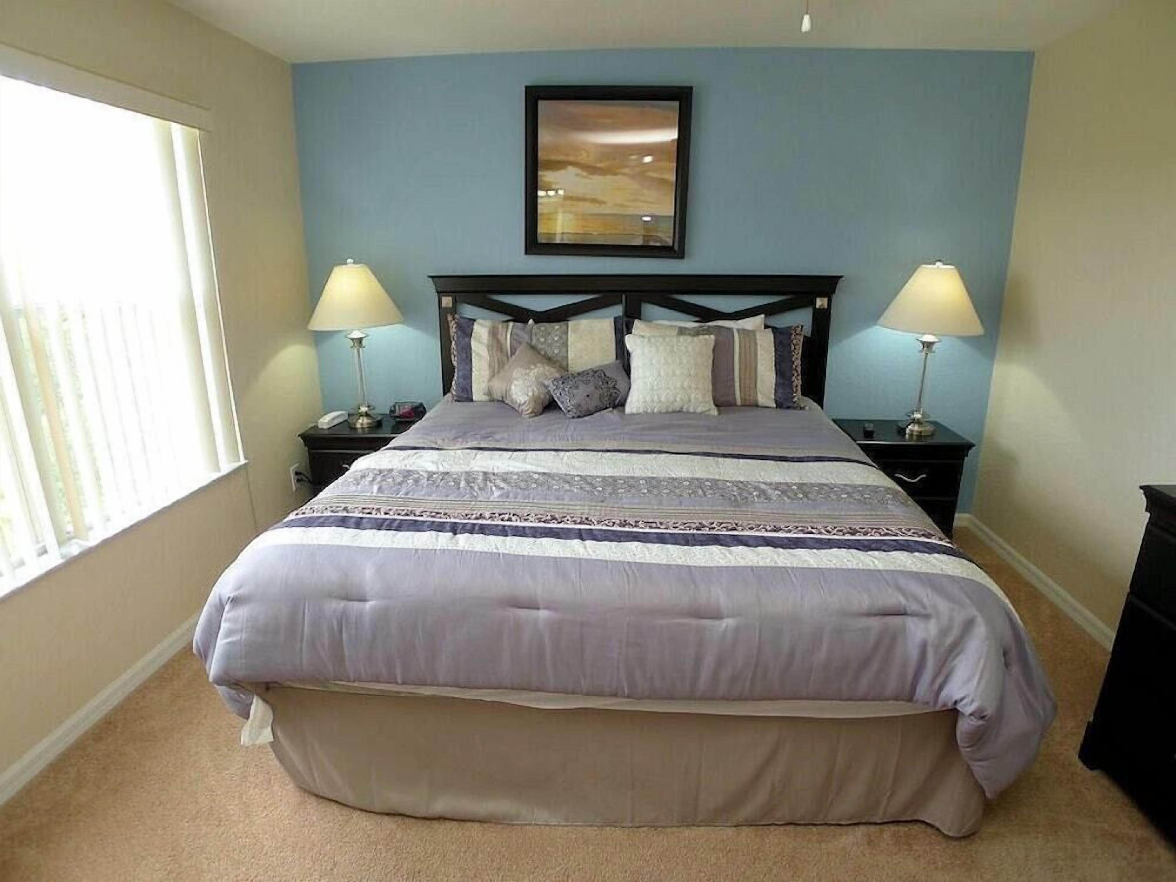 A bedroom with a large bed, two bedside tables with lamps, a framed picture above the bed, and a window on the left.