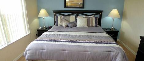 A bedroom with a large bed, two bedside tables with lamps, a framed picture above the bed, and a window on the left.
