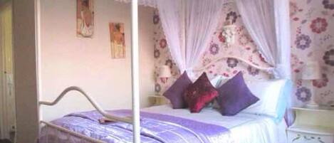 Quarto com cama de dossel, papel de parede floral e mesa de cabeceira.