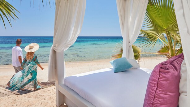 A beachside room with a bed, white curtains, and a view of the ocean.