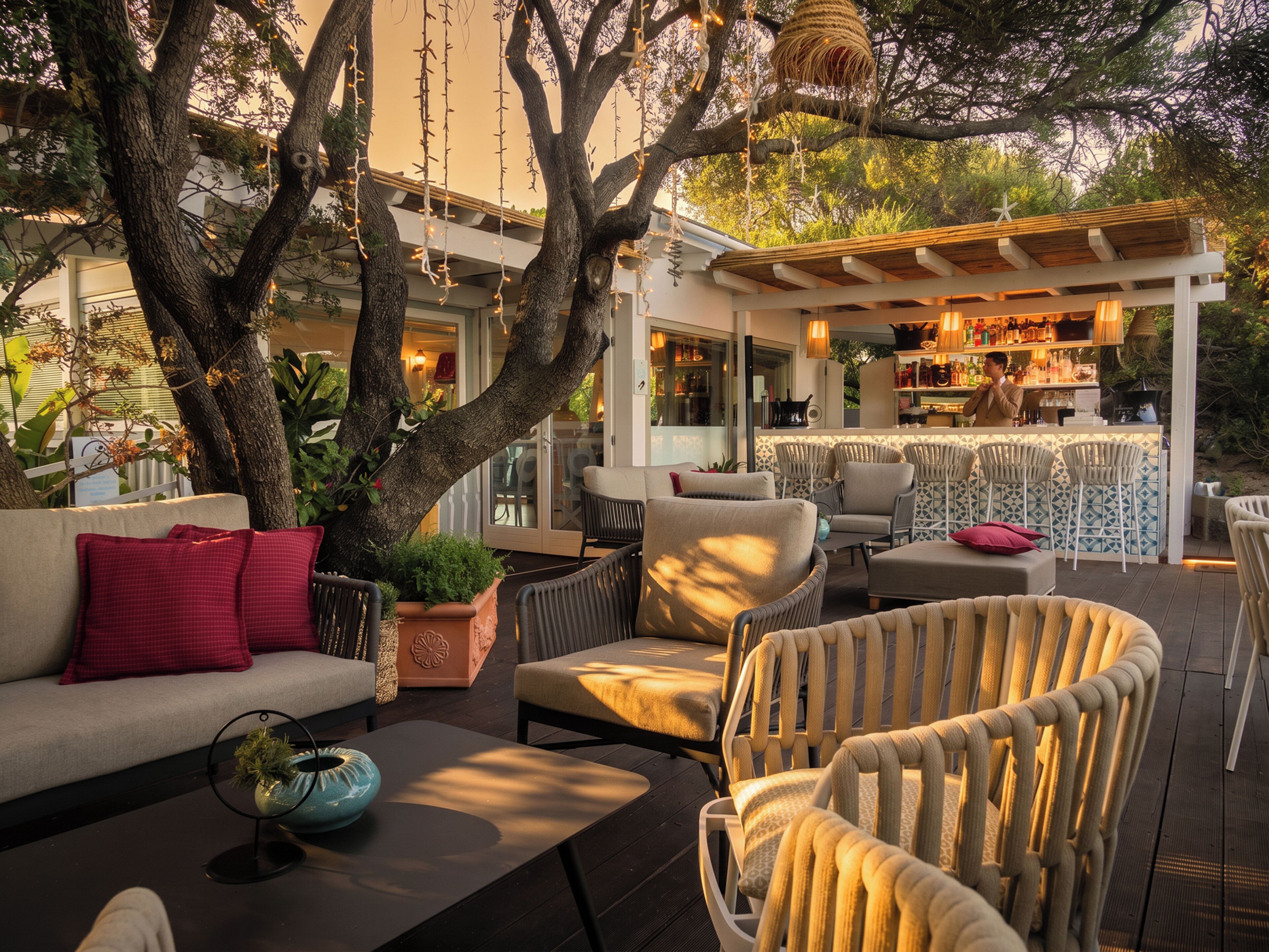 An outdoor seating area with a bar, cushioned chairs, a sofa, and a large tree.