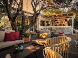 An outdoor seating area with a bar, cushioned chairs, a sofa, and a large tree. - Forte Village Resort - Le Dune (Pula)