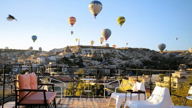 En terrasse med udendørs siddepladser og udsigt til varmluftsballoner over et bybillede.