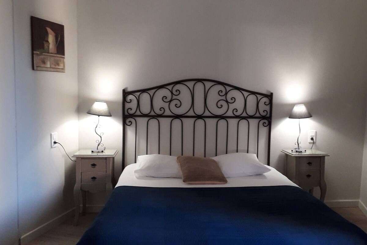 A neatly made bed with a blue bedspread and a brown pillow, flanked by two bedside tables with lamps, in a room with plain walls and a simple metal headboard.