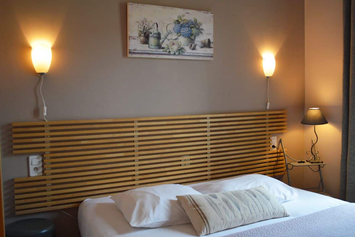 A hotel room with a wooden headboard, a bed with white linens, two wall-mounted lamps, and a painting above the headboard.