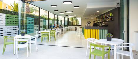 Un área de estar al aire libre con sillas blancas y verdes, un bar con estantes llenos de botellas y un muro de cristal con vistas a un jardín.