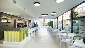 Uma área de bar moderna com um balcão comprido, cadeiras e mesas brancas. Garrafas estão expostas em prateleiras e uma pessoa trabalha atrás do balcão.
