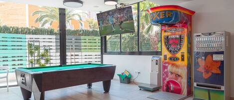 Una mesa de billar, una pantalla grande y una máquina recreativa colorida en una habitación con ventanas amplias.