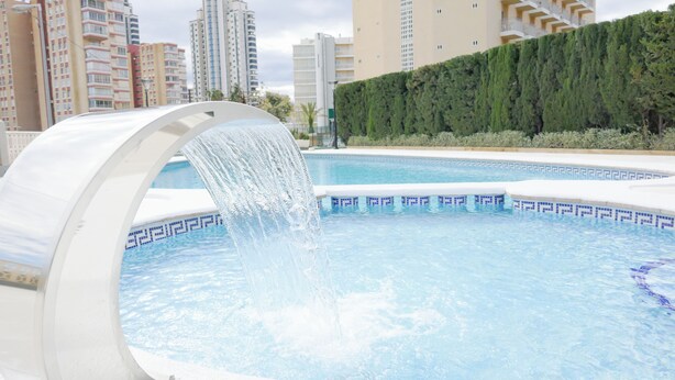 Piscina com um elemento aquático, cercada por edifícios altos e um sebedo.