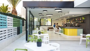 Um café ao ar livre com mesas brancas e cadeiras verdes, uma área de bar com balcão amarelo e vista para palmeiras e edifícios ao fundo.