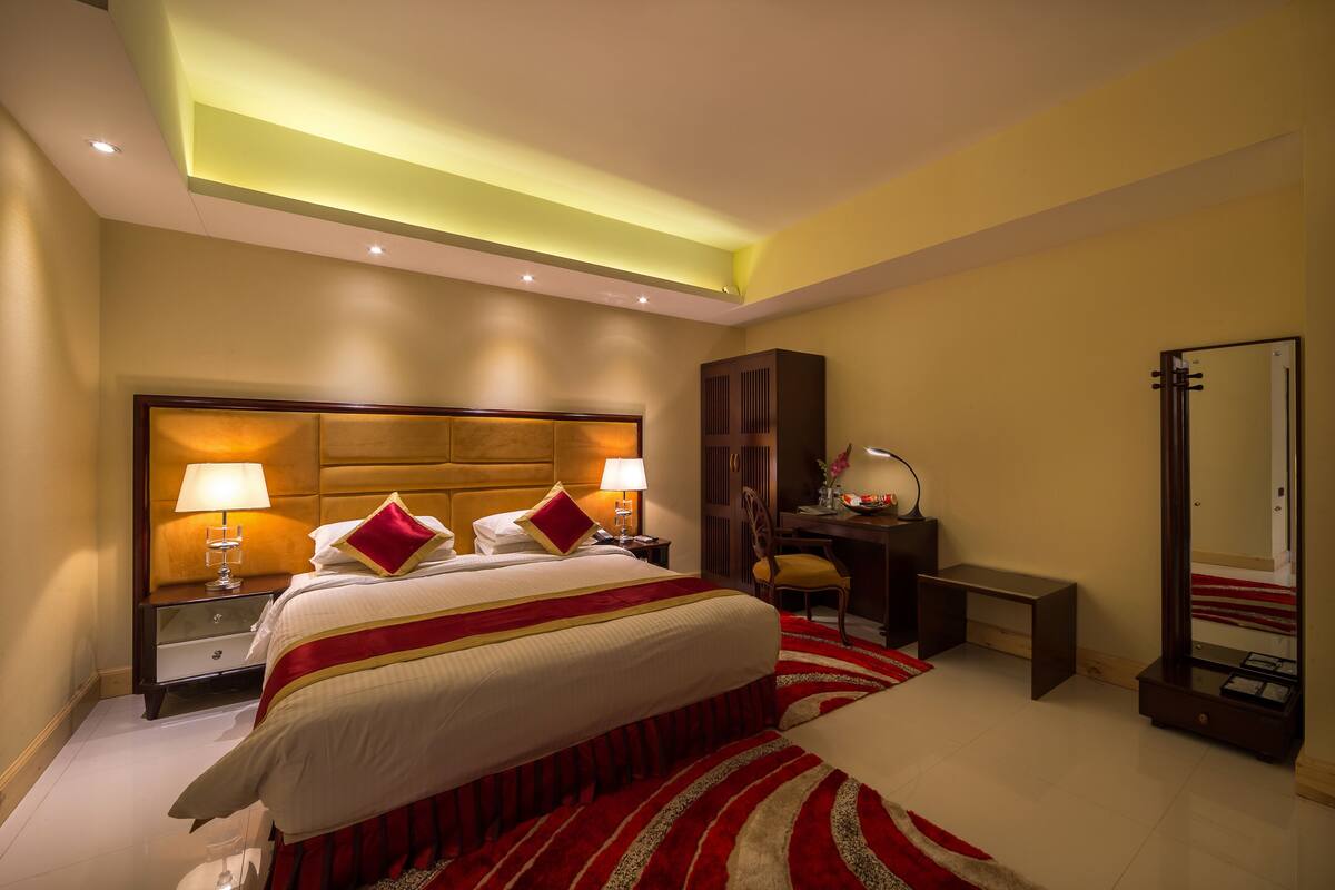 Una habitación de hotel con una cama grande, lámparas de noche, un escritorio, un espejo y una alfombra a rayas rojas y blancas.