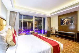 Habitación de hotel con una cama grande, televisión, balcón con vistas y zona de estar con mesa y sillas.