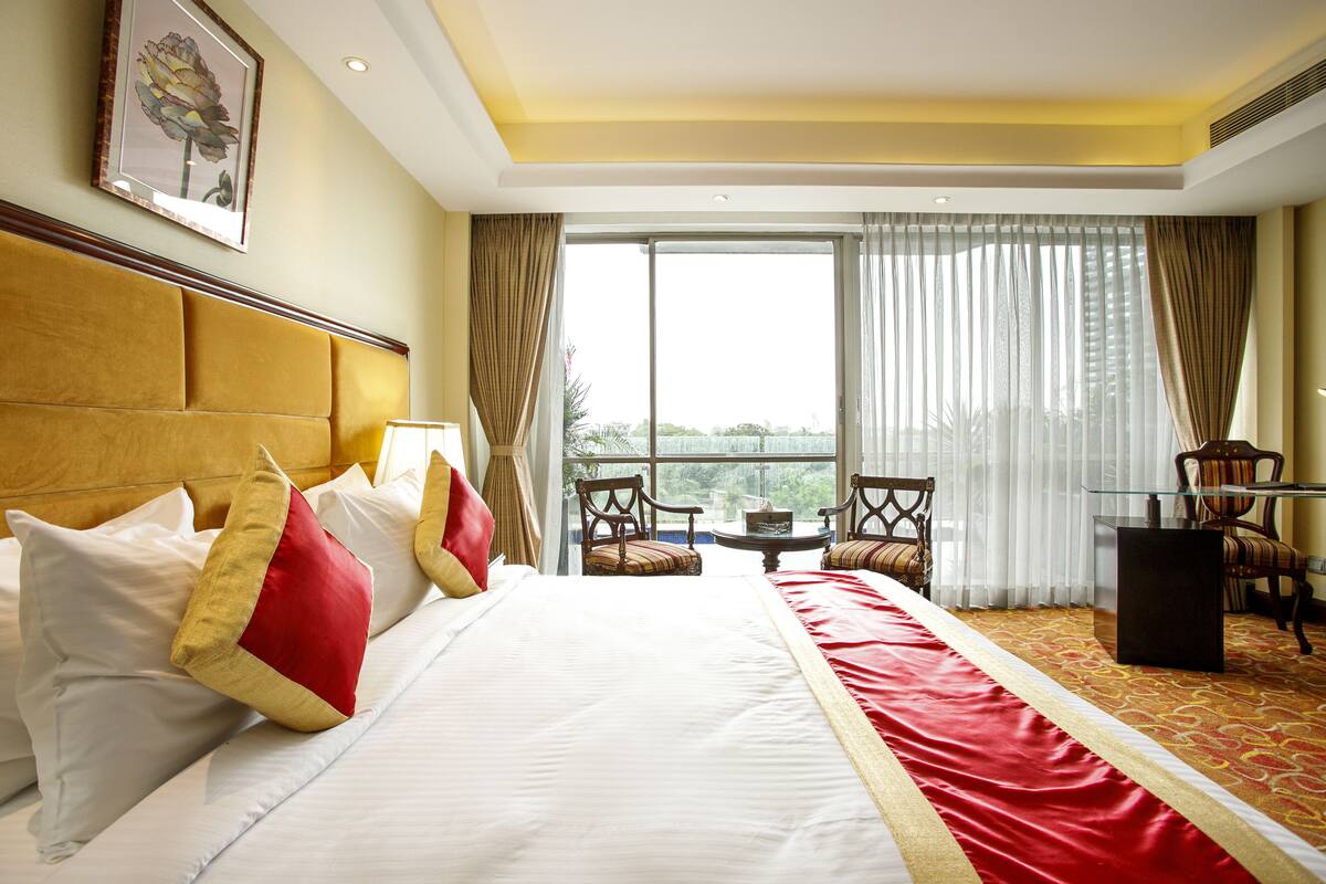 Habitación de hotel con una cama grande, un corredor de cama rojo y blanco, un cabecero de madera, dos sillas, una mesita y vistas a zonas verdes por la ventana.