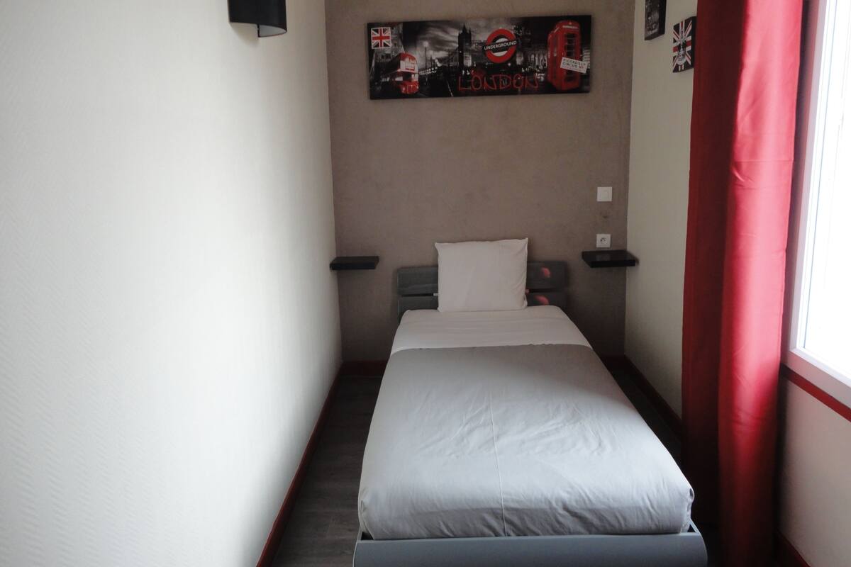 Een hotelkamer met een bed, nachtkastje, een wandlamp en een poster van bezienswaardigheden in Londen.
