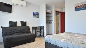 Une chambre moderne avec un canapé noir, un lit recouvert d’une literie à motifs floraux, un bureau et un tableau représentant un paysage urbain.