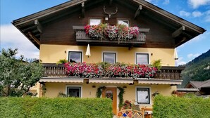 Un chalet de dos pisos con un balcón adornado con flores, fachada de madera y montañas al fondo.