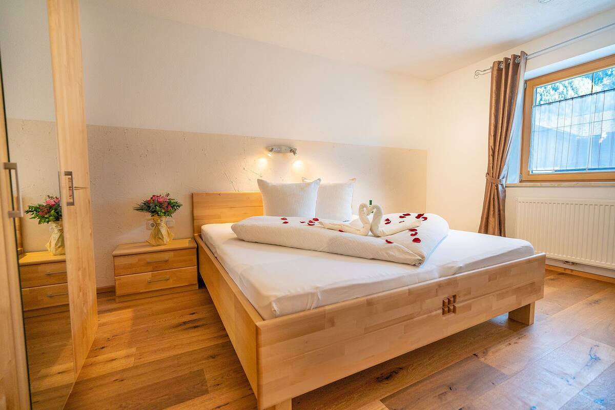 A bedroom with a wooden bed, bedside tables, a nightstand with a vase of flowers, and a window with curtains.